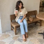 Woman seated in a café wearing a beige linen shirt, ivory tank, light blue jeans, and tan Tabi flats beside a small shoulder bag.
